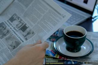 a person reading a newspaper with a cup of coffee by Anna Keibalo courtesy of Unsplash.