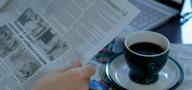 a person reading a newspaper with a cup of coffee by Anna Keibalo courtesy of Unsplash.